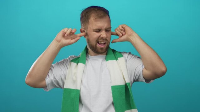Young caucasian man plugging his ears with fingers while wearing a green and white soccer scarf in a turquoise studio; frustration.