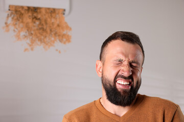 Man grimacing because of bad smell from air conditioner indoors. Dirty air flows from appliance