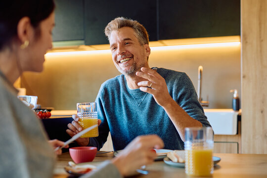 Happy man and woman having breakfast together, laughing while drinking orange juice and talking in a modern kitchen