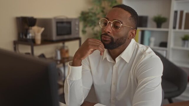 Man wearing white shirt and round glasses rests chin on hand at office desk while studying a monitor; pensive concentration.