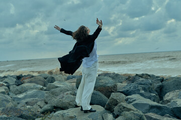 Two people on a rocky shore share a joyful moment, arms raised, wind in their coats, sea breeze and cloudy sky celebrating freedom and outdoor adventure
