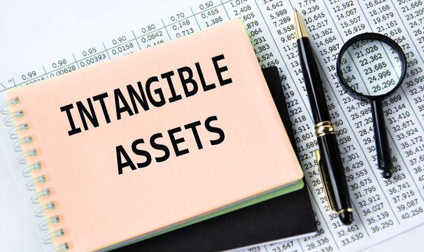 INTANGIBLE ASSETS - words on a yellow notebook on a background of digital tables, a magnifying glass and a pen