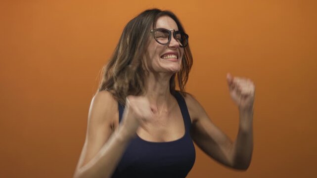 Woman wearing black glasses with clenched fists and a wide grin stands on an orange studio set; excitement.