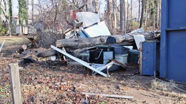 Shed is damaged surrounded by trash discarded objects in wooded area with bare trees.