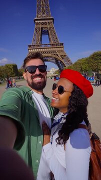 Vertical. Happy interracial couple taking a selfie in Paris with the Eiffel Tower behind. Casual travel portrait on the grass with sunglasses and beret, conveying joy, romance and city vibes.
