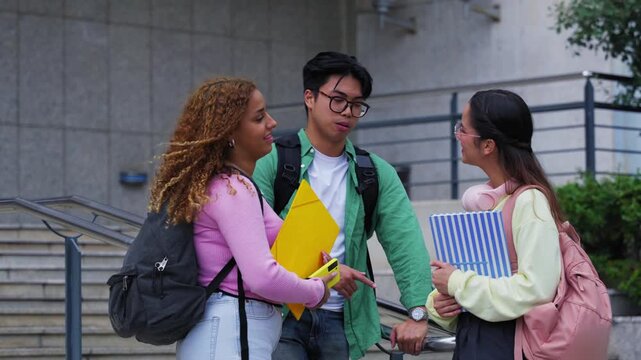 Three university students talk on campus steps after class, holding folders and backpacks. Casual mood, diverse group, one student glances at camera while friends smile during a short break.