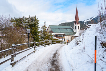 Fenced snowy path leading to a mountain village in Tyrol on a winter day. A traditional church with a tall bell tower is visible in background.