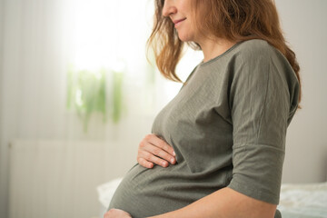 Serene expecting mother sitting on bed, lovingly caressing her belly in cozy home setting. Beautiful moment of maternal connection and anticipation.