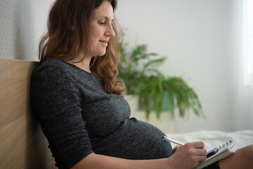 Pregnant woman holding pen and notebook, preparing a birth plan while sitting on bed.