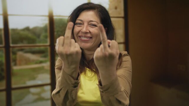 Woman in yellow shirt shows two middle finger gestures in room with window grid and sunlight streaming through; defiance.