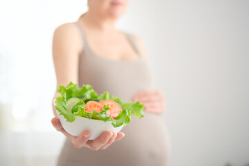 Expecting mother holding fresh vegetable salad while gently touching her belly. Healthy nutrition moment during pregnancy in cozy home setting.