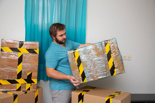 Mature man with beard looks at picked up cardboard box with yellow and black tape in empty room of apartment. Concept of moving, immigration, starting new life, domestic life, rent apartment, mortgage