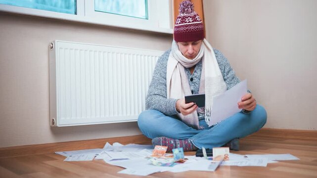 Stressed man in winter clothes sits by radiator checking high heating bills. He crumples paper in anger and covers face in despair over financial crisis and inflation. High quality 4k footage