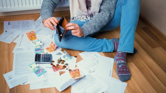 Man in warm clothes sits on floor among scattered bills and shakes an empty wallet upside down. Concept of poverty, bankruptcy, and rising heating costs during financial crisis.High quality 4k footage