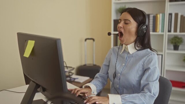 Woman yawning and stretching with headset at computer monitor in home office building with rolling suitcase and bookshelf visible; fatigue.