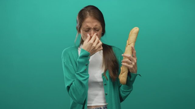 Young hispanic woman pinched nose while holding baguette in teal studio reacting to an unpleasant smell; disgust.