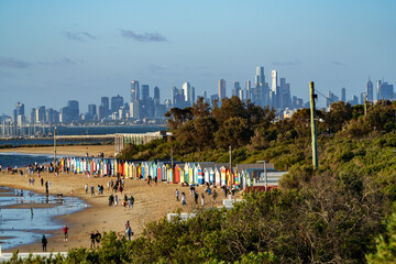 Fototapeta premium Vibrant bathing boxes line Brighton Beach, filled with people, overlooking the calm bay and Melbourne's impressive city skyline under a clear sky.
