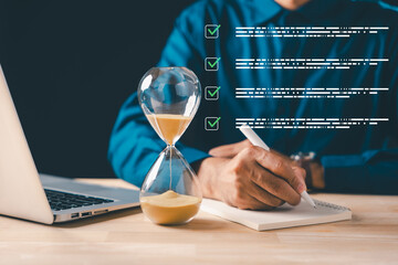 Business professional using pen to write notes in book, hourglass showing passing time, laptop...