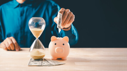 Hand places money into piggy bank, hourglass shows passage of time, finance concept for smart saving and investment promoting financial growth for future purchase and stock market activity