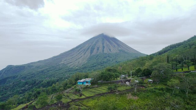 Cinematic drone forward shot of majestic Mount Inerie volcano with a cloudy peak, overlooking a rural village and green forest in Flores, Indonesia.