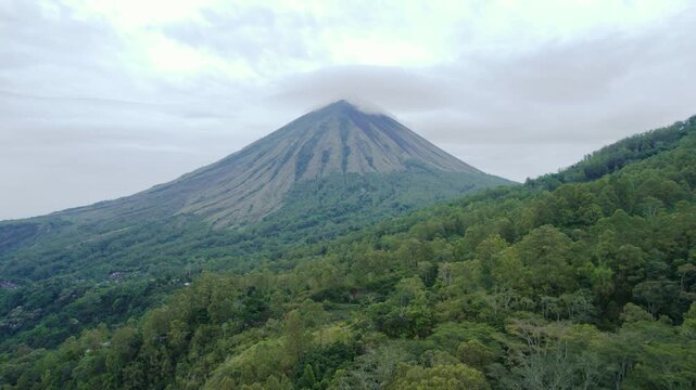 Cinematic drone forward motion of Mount Inerie volcano under cloudy sky, surrounded by dense green forest in Flores, Indonesia.