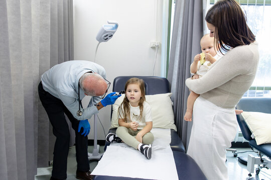 Doctor checking young girls ear with otoscope with mum carrying toddler son beside