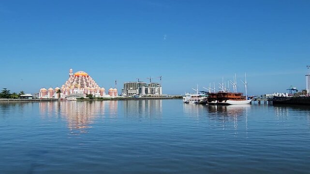 A scenic waterfront view of the 99 Domes Mosque (Masjid 99 Kubah) and traditional Phinisi schooner boats reflected in the calm waters of Losari Beach under a clear blue sky.