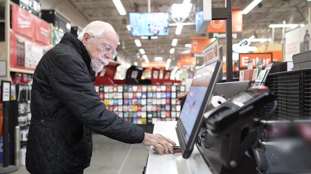 An elderly man has trouble using the self checkout system while shopping at a hardware store during peak hours.