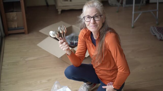 A mature woman examines silver cutlery pieces, recalling family memories and sentiments tied to inherited heirlooms in her home.