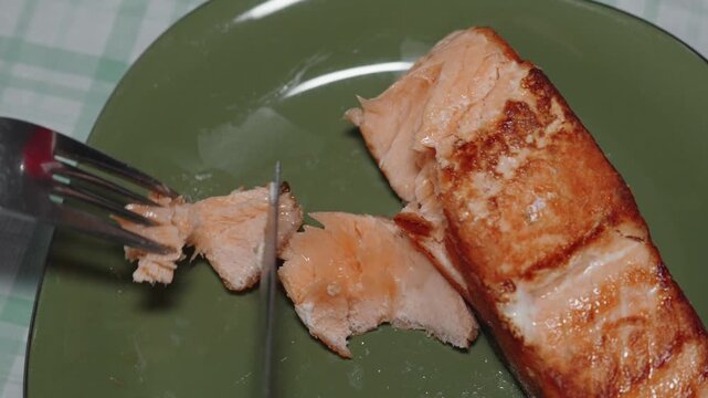 Close-up of a fork and knife breaking apart pieces of grilled salmon on a green plate, representing healthy eating and omega-3 rich food