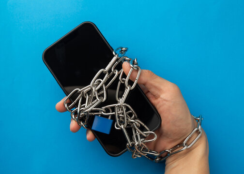 Top view of a hand chained to a smartphone with a blue padlock. A metaphor for social media addiction, digital dependency, technology trap, and the importance of a digital detox lifestyle. - Powered by Adobe