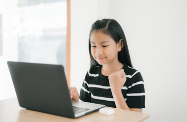 A young girl is sitting at a table, concentrating as she uses a laptop for studying or online learning in a bright, calm home environment.