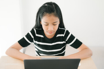 A young girl is sitting at a table, concentrating as she uses a laptop for studying or online learning in a bright, calm home environment.