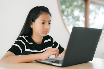 A young girl is sitting at a table, concentrating as she uses a laptop for studying or online learning in a bright, calm home environment.