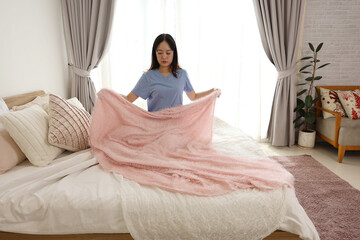 Diligent young asian woman making bed and holding soft pink blanket in cozy bedroom interior organizing home with focused and attentive expression during morning routine at house apartment