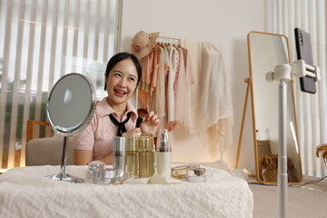 Young smiling asian woman influencer recording video tutorial with smartphone creates beauty content for social media audience featuring cosmetic skincare product review in modern bedroom studio