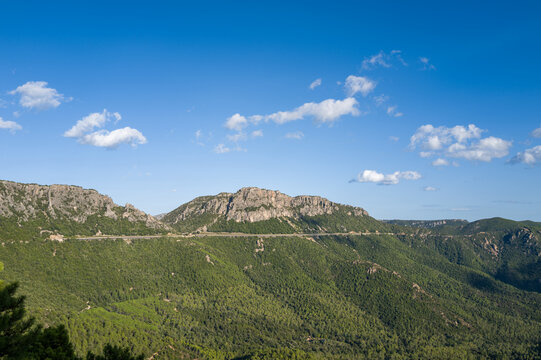 A long elevated road slices across a vast expanse of green forest and rugged mountains near Baunei and Dorgali in Sardinia, under a vivid blue sky with scattered clouds.