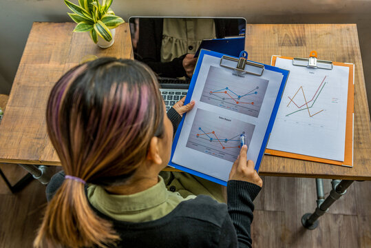 Woman reviewing business charts and graphs on clipboards at wooden desk, analyzing company performance metrics, sales growth trends and financial data in modern home office environment

