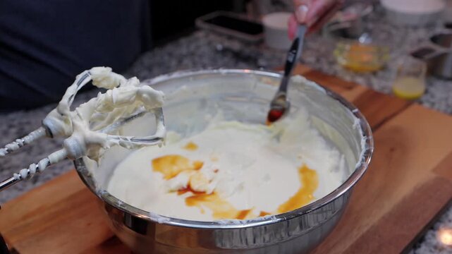 person pouring vanilla extract from a measuring spoon into cheesecake batter in a metal bowl