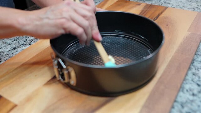 person greasing a black springform cake pan with butter using a silicone brush on a wooden board