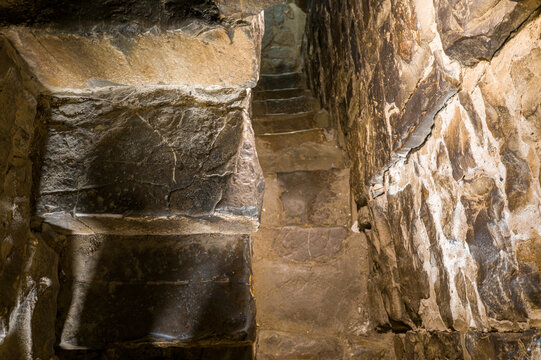 Rough-hewn stone staircase ascends through a confined passageway, with textured masonry walls illuminated by soft natural light. The scene evokes a sense of age, mystery, and medieval architecture.