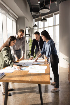 Diverse team of business analytic professionals reviewing statistic repots together, bending over table with papers, discussing marketing research results, developing strategy of growth. Vertical shot