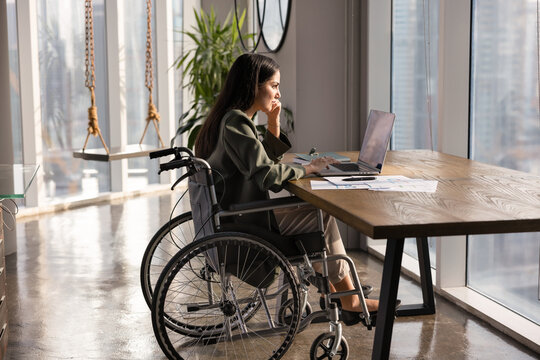 Focused young Latin businesswoman with disability working on Internet project in modern office, typing on laptop at paper marketing reports, sitting at workplace table in wheelchair