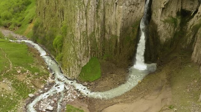 An aerial view captures a stunning, narrow waterfall cascading down a sheer, rocky cliff face in the remote and rugged landscape of the Kara-Syrt Ridge. The powerful flow of water meets a winding rive