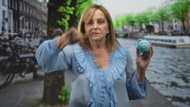 Middleage woman points finger while holding blue piggy bank on a street by a canal; financial caution.
