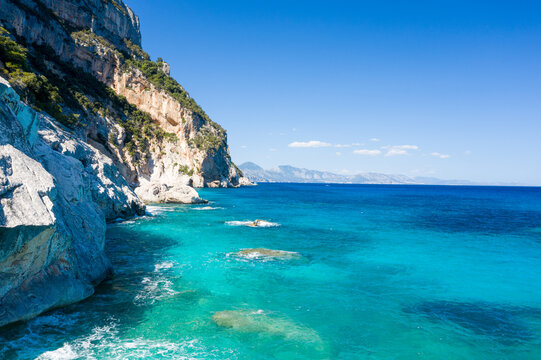Steep limestone cliffs rise above crystal-clear turquoise water along the rugged coastline of Cala Goloritze, Sardinia. Bright sunlight and a vivid blue sky create a striking, serene Mediterranean