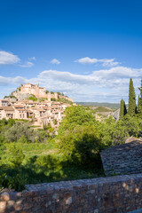 Fototapeta premium Elevated view of Alquezar, Spain, featuring a prominent medieval fortress above terracotta-roofed houses, lush greenery, and cypress trees under a vibrant blue sky with scattered clouds.