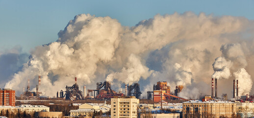 Factories release large clouds of smoke into the air while buildings sit at the forefront. This...