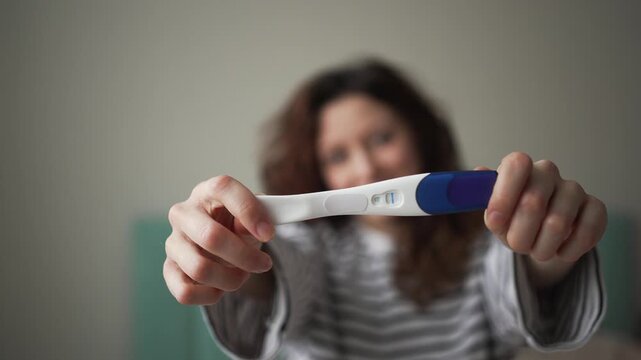 Out-of-focus woman smiling and happily showing a positive pregnancy test with two lines, bringing it into focus towards the camera, celebrating the beginning of a new chapter in her life