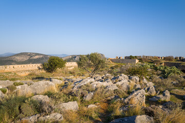 Weathered stone fortifications stretch across a rugged hillside dotted with rocks, dry grass, and sparse shrubs at Palamidi Fortress in Nafplio, Greece. Warm sunlight and clear blue sky create a